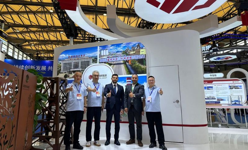 Four men, three in matching polo shirts and one in a suit, stand in an exhibition stand giving thumbs-up. The stand has banners, images of buildings, and informational displays under a bright yellow ceiling.