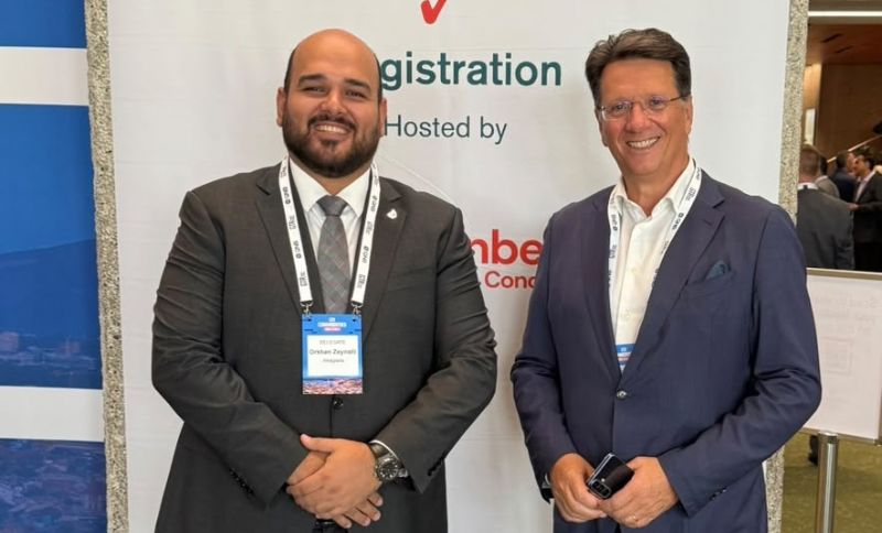 Two men in business attire stand smiling in front of a registration sign at a conference event. Both wear suits and conference badges around their necks.