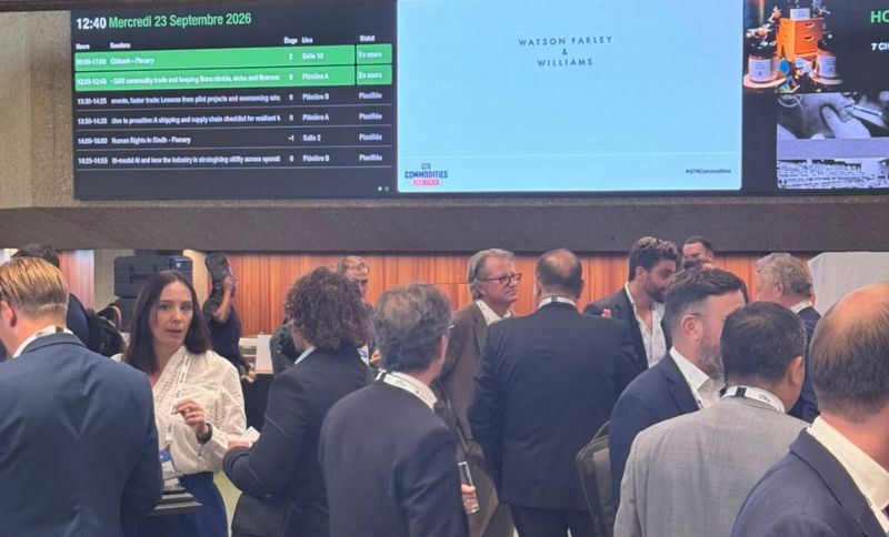 People in business attire converse at an indoor networking event. Above them, a large screen displays an event timetable and logos. The atmosphere is busy and professional.