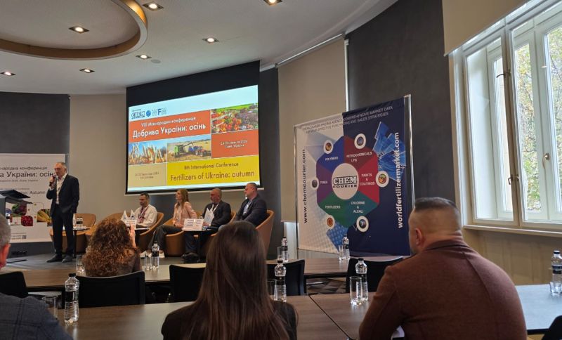 A conference room with five panellists seated on stage, a speaker standing, and an audience watching. A presentation screen displays information about fertilisers and Ukrainian culture. Large banners and windows are visible.