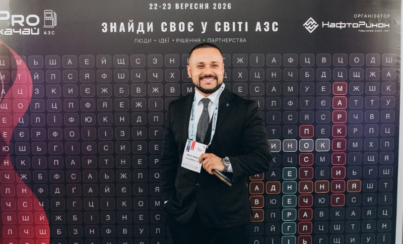 A man in a suit smiles at the camera, holding a badge, standing in front of a backdrop with Cyrillic letters and Ukrainian text promoting a September 2026 event.