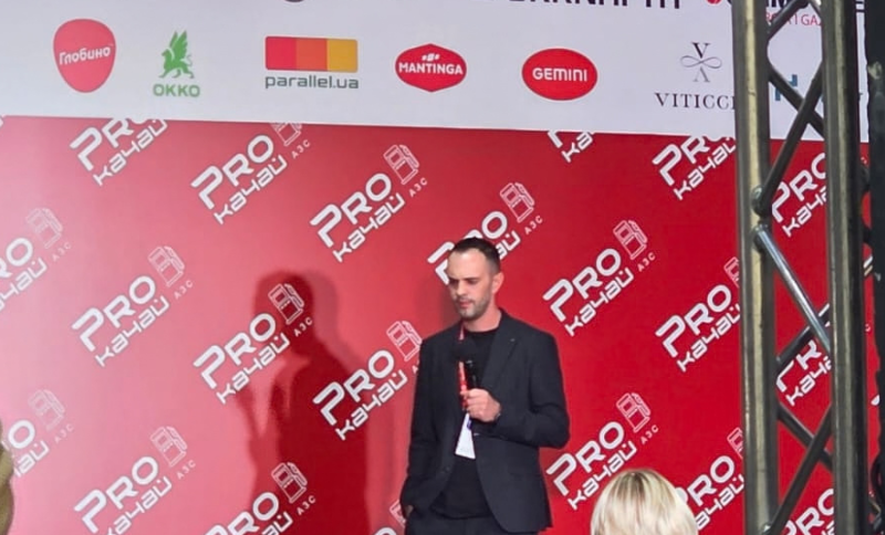 A man in a black suit speaks on a red stage with Pro Kašteli logos, holding a microphone. Sponsor logos are displayed above him on a white banner. An audience member is visible in the foreground.