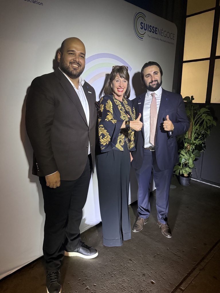 Three people in business attire stand smiling and giving thumbs up in front of a Suisse Negoce backdrop, with plants and a window to the right.