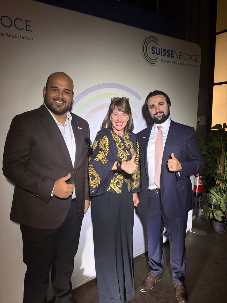 Three people dressed in business attire stand smiling and giving thumbs up in front of a SUISSENÉGOCE event backdrop.