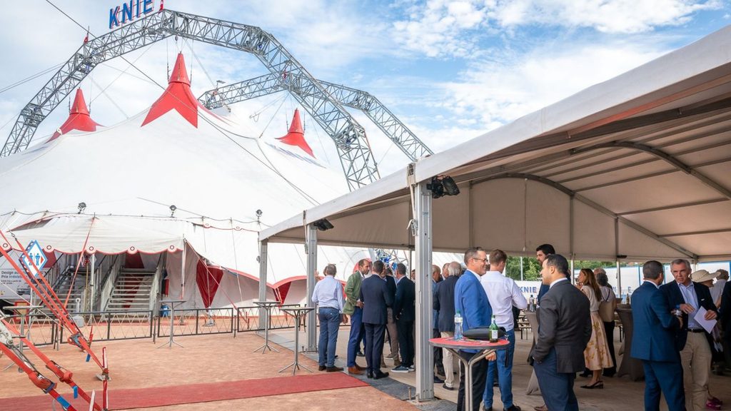 A group of people in formal attire gather outside a large white circus tent with red peaks under a partly cloudy sky. The tent has KNIE signage above, and a covered area with more guests is nearby.