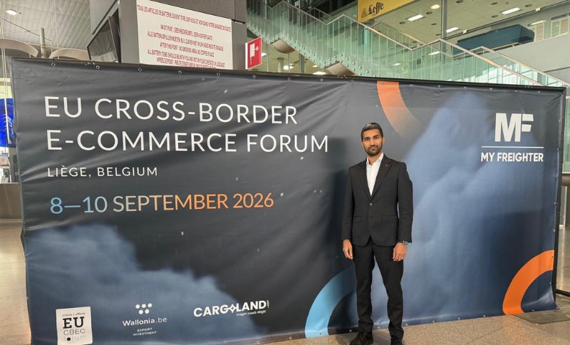 A man in a black suit stands next to a banner for the EU Cross-Border E-Commerce Forum in Liège, Belgium, taking place from 8–10 September 2026. The banner features logos and sponsor names.