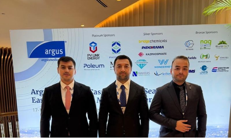 Three men in suits stand in front of a sponsor banner at an event. The banner displays various company logos under Platinum, Silver, and Bronze Sponsor categories. Warm lighting and modern décor are visible in the background.