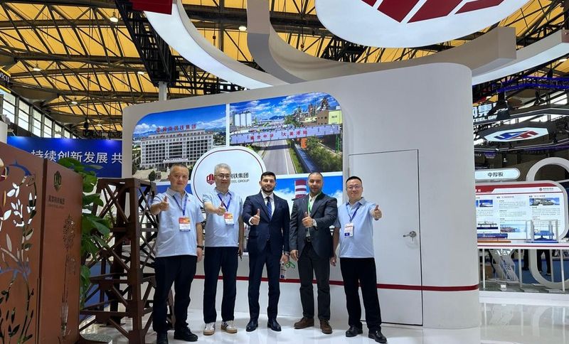 Four men, two in suits and two in white shirts, stand indoors at an exhibition stand, giving thumbs up. Display boards and a large curved structure are visible behind them. The scene appears professional and friendly.