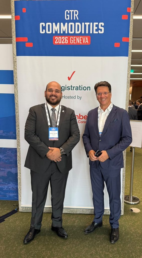 Two men in business attire stand smiling in front of a GTR Commodities 2026 Geneva event banner at a conference. Both wear badges around their necks.