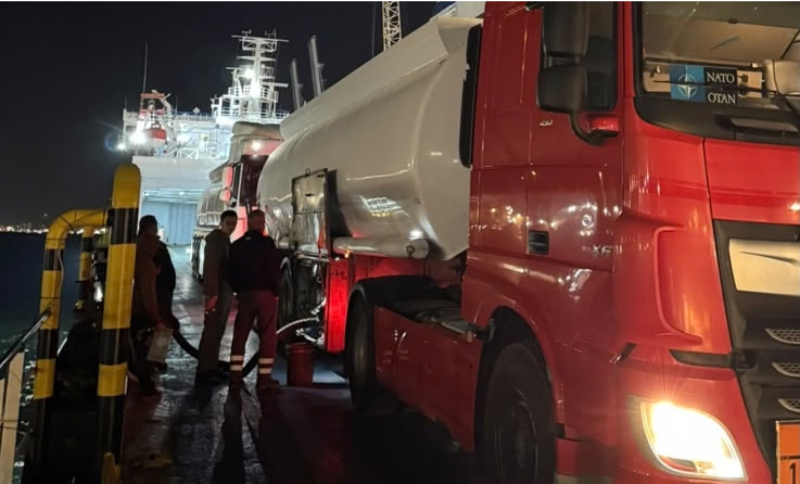 A red tanker lorry parked on a ship deck at night, with two people attending to a hose connection near the tank.