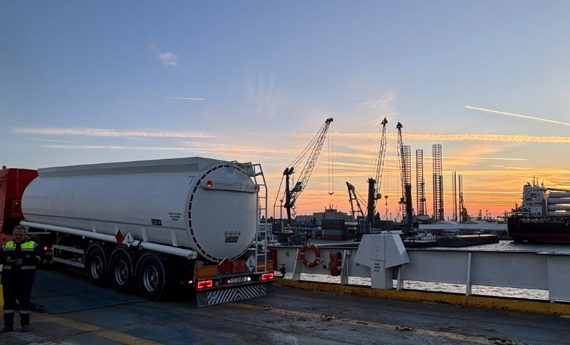 A tanker lorry parked at a shipping terminal at sunset, with port cranes and cargo ships in the background.