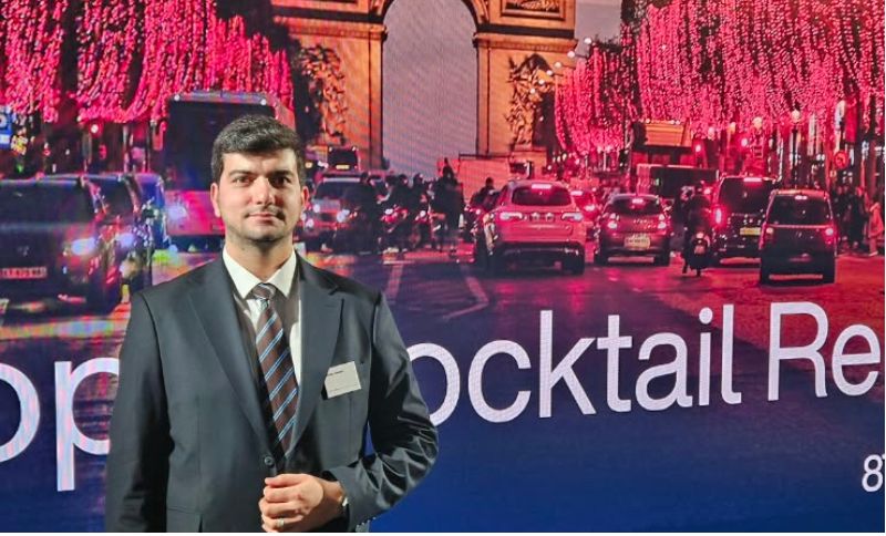 A man in a suit and tie standing in front of a digital screen displaying a nighttime street scene in Paris with the words Cocktail Reception.