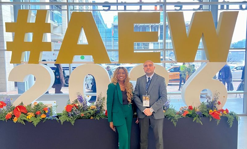 A man and a woman posing in front of a large gold #AEW 2026 sign.