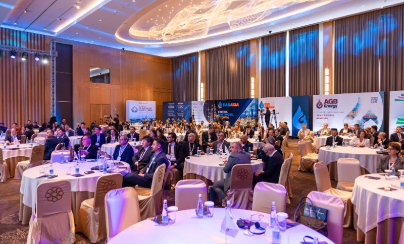 Attendees sit at round tables in a large conference hall with branding displays for energy companies in the background.