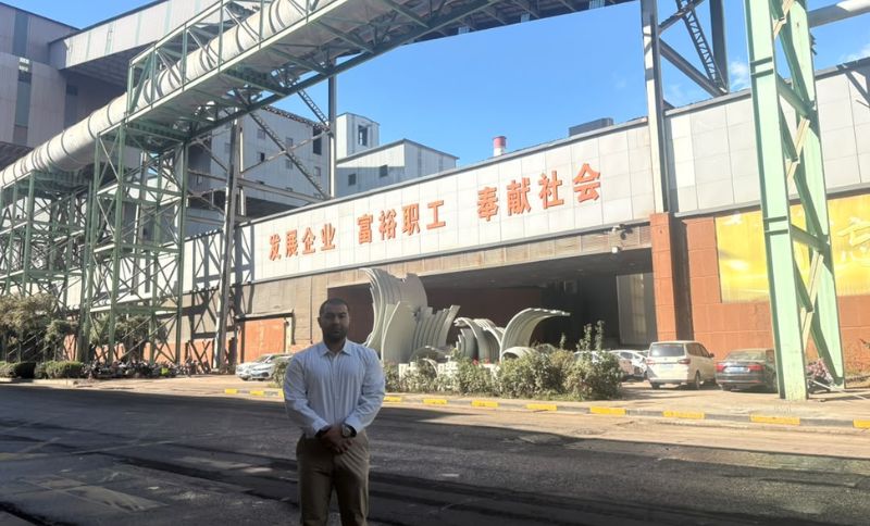 A man stands in front of a large industrial building with orange Chinese characters displayed above the entrance.