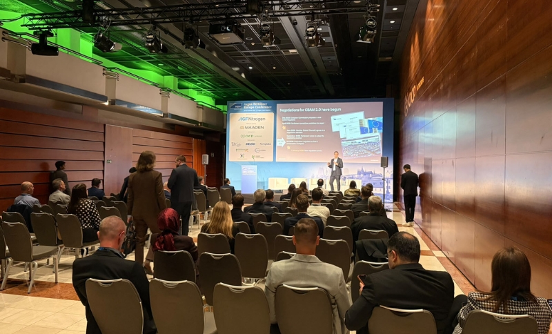 A speaker stands on a stage before an audience in a conference hall, presenting in front of a large screen displaying charts and text about CBAM 2.0.