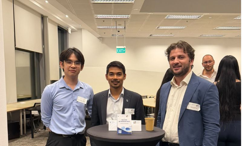 Three men in business attire stand behind a small table with a networking event sign in an office setting.