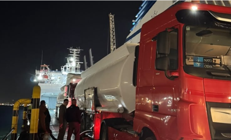 A red lorry with a white tanker trailer parked on the deck of a ferry at night, with three people standing near the vehicle.