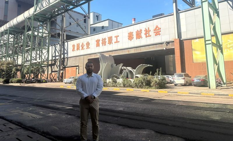 A man in a white dress shirt and tan trousers stands in front of a large industrial building with red Chinese characters above the entrance.