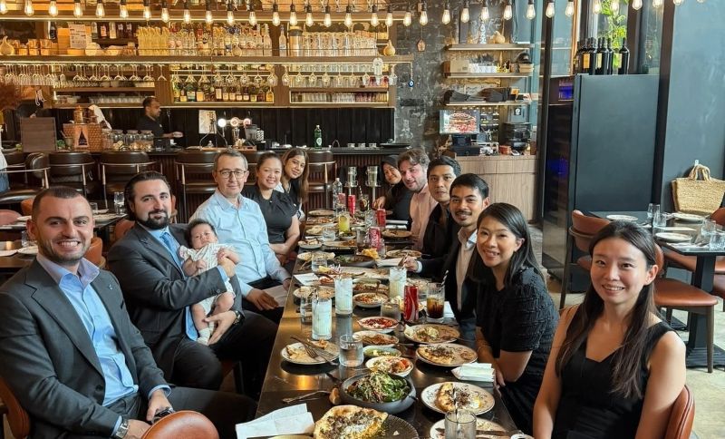 A group of people sitting at a long dining table in a restaurant with a bar in the background.