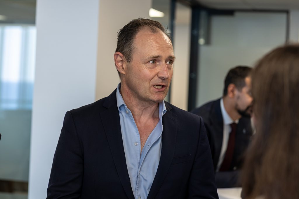 A man in a navy blazer and light blue shirt speaks to a person whose back is to the camera.