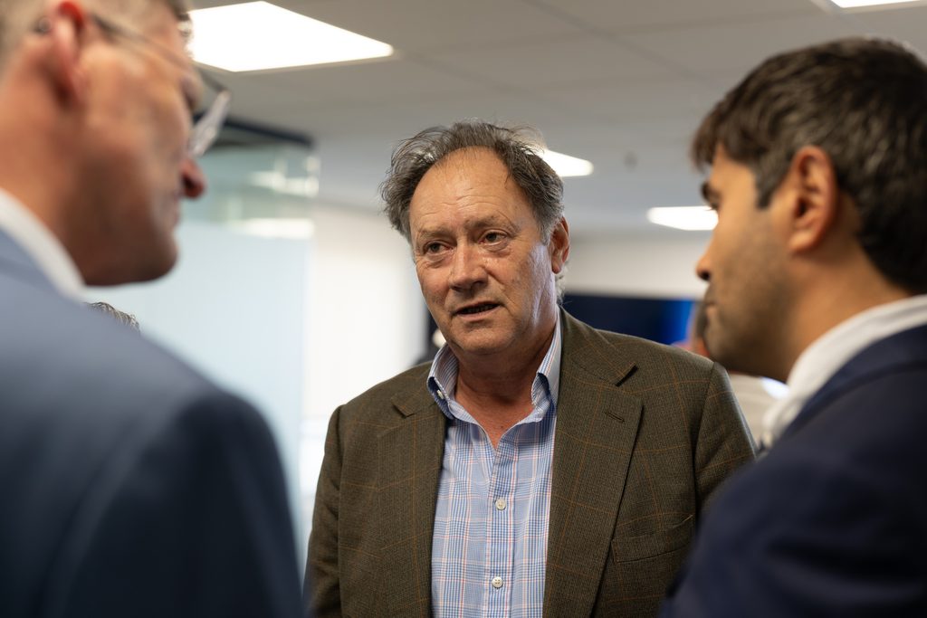 A middle-aged man in a checked blazer and light blue button-down shirt speaks to two other men in suits in an office setting.