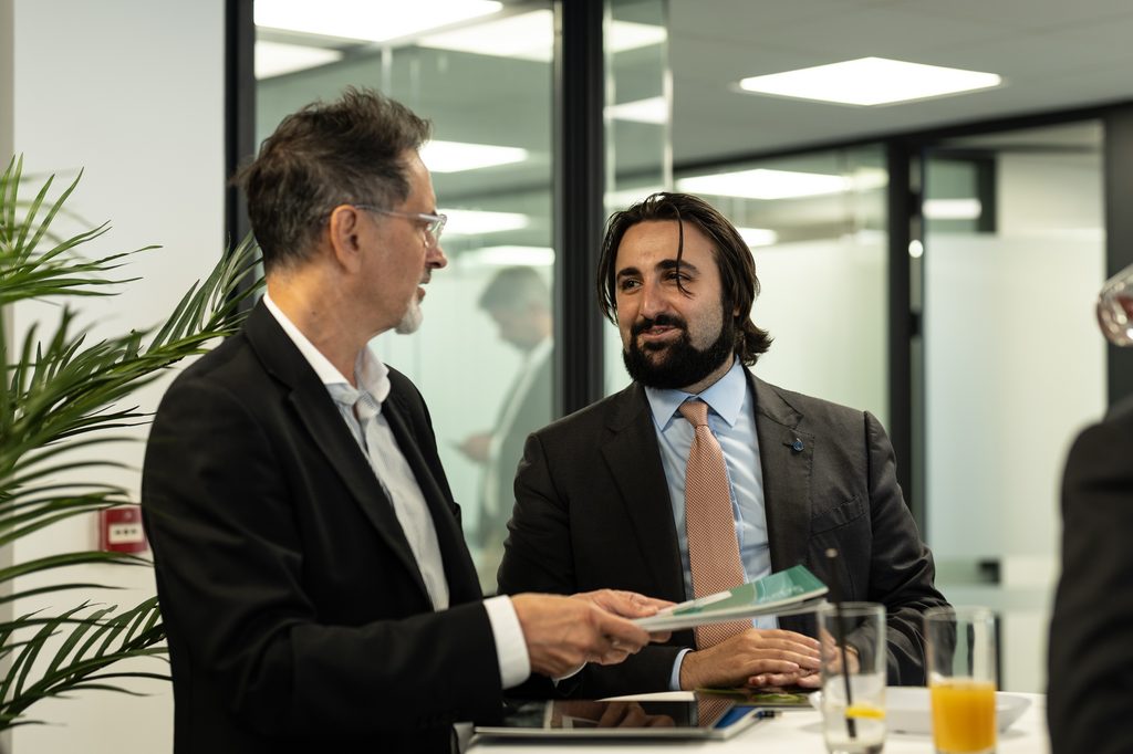 Two men in business suits talking and holding a leaflet at a networking event.