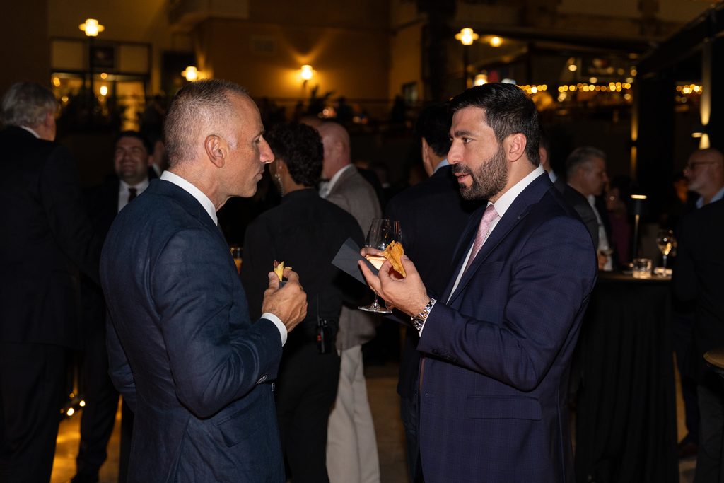 Two men in suits talk whilst holding canapés and drinks at an outdoor evening social event.