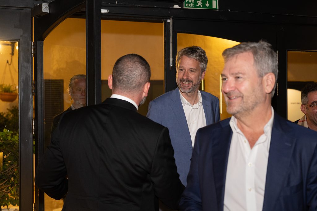 Three men in business attire stand near a doorway at an evening event.
