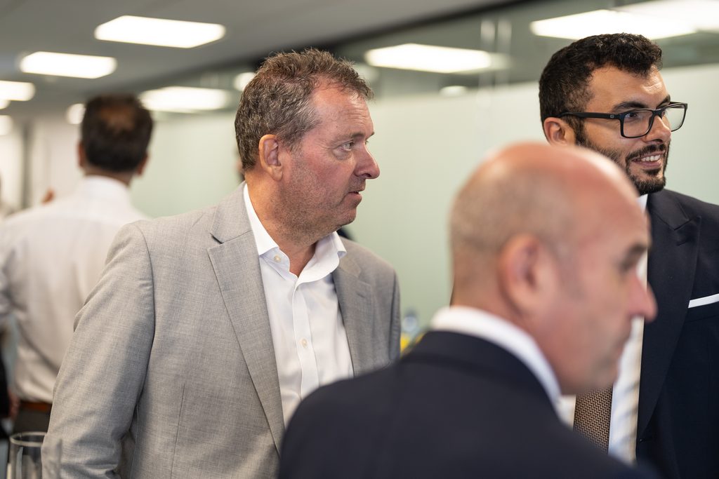 Three men in business attire stand in an office setting, with two of them engaged in conversation.