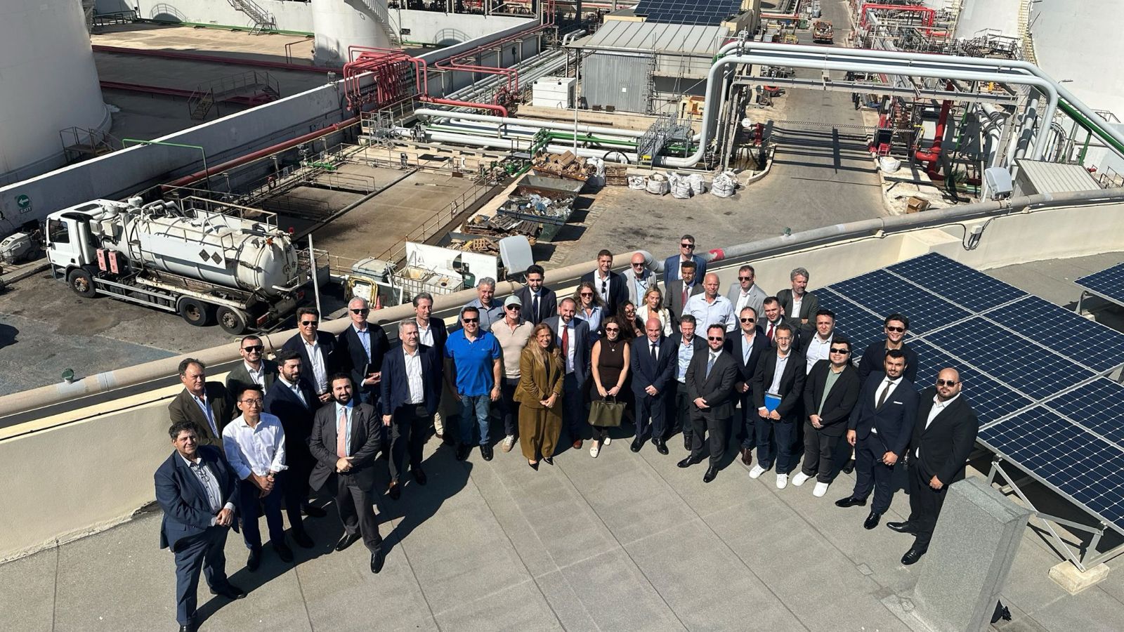 A large group of people in business attire posing for a photo on a rooftop terrace overlooking an industrial facility with solar panels and storage tanks.