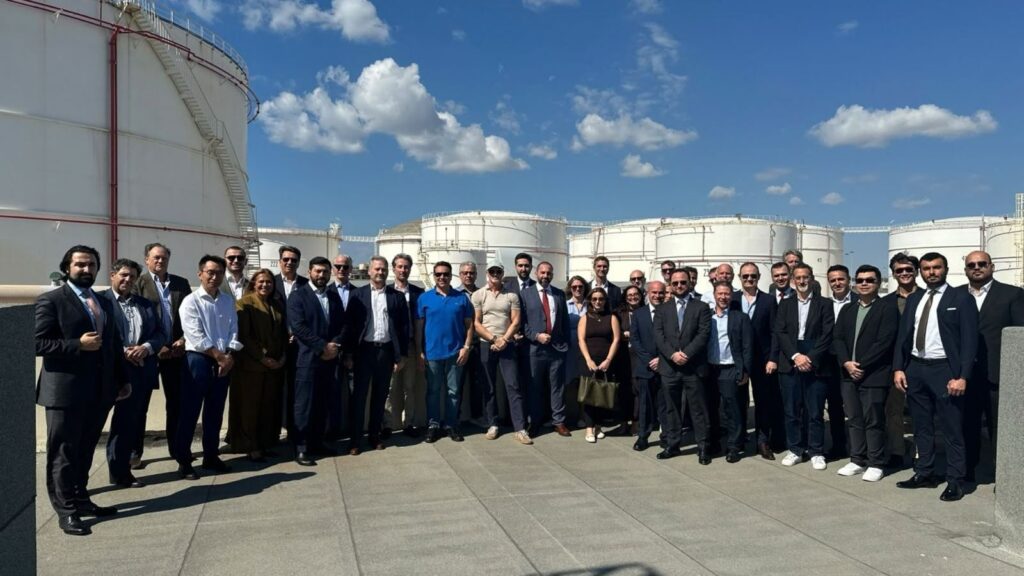 A large group of professional men and women posing for a photo on a rooftop platform in front of several industrial storage tanks.