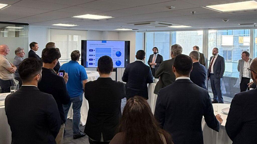 A presenter stands before a group of people in a meeting room, pointing to a large screen displaying a chart titled Commodities Coverage.