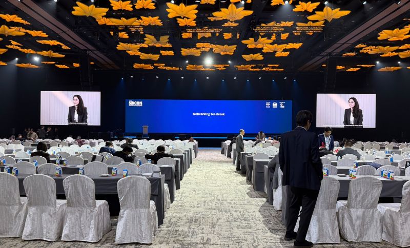 A large conference hall with rows of tables and white-covered chairs facing a stage with a blue screen reading Networking Tea Break, featuring large yellow flower-shaped ceiling decorations and two side screens displaying a speaker.