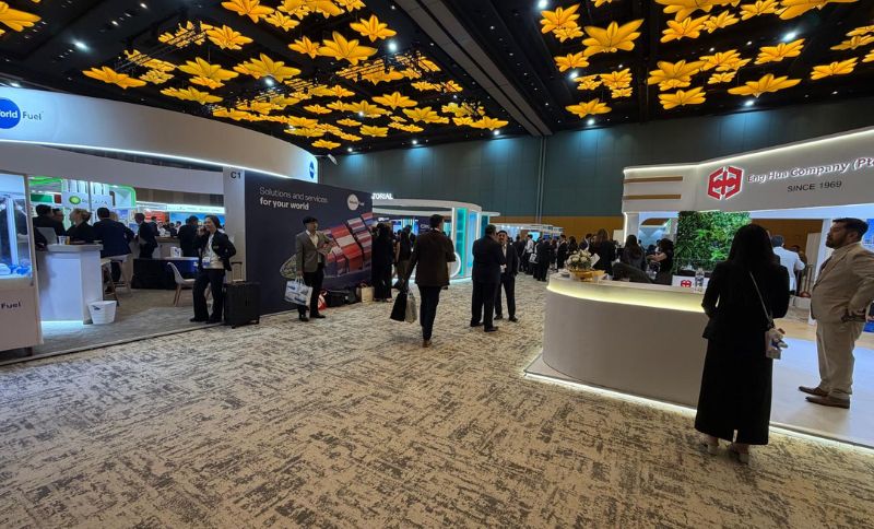 People walk through a large trade show exhibition hall featuring company stands and a ceiling decorated with yellow floral-shaped light fittings.