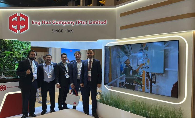 Five men in business attire stand in front of an Eng Hua Company trade show stand featuring a large screen displaying an industrial worker operating machinery.